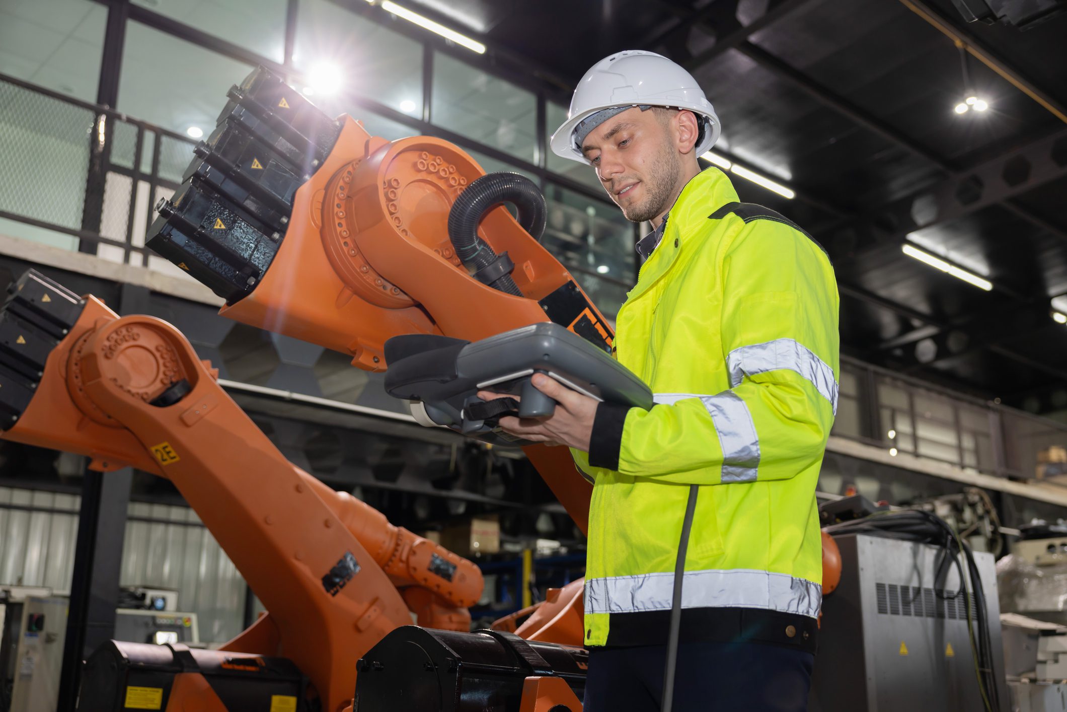 Caucasian worker in a bright yellow safety jacket and helmet controls an industrial robot arm in a factory