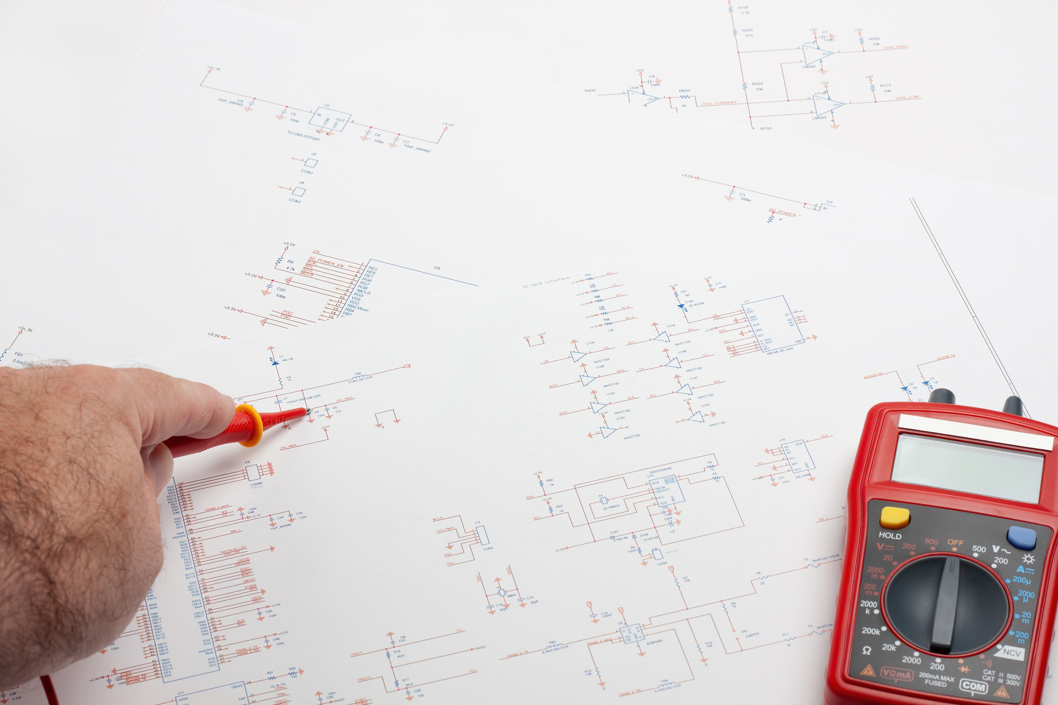 An engineer's desk featuring a hand holding a measuring cord over electronic blueprints. The image highlights the process of designing and reviewing electronic components.